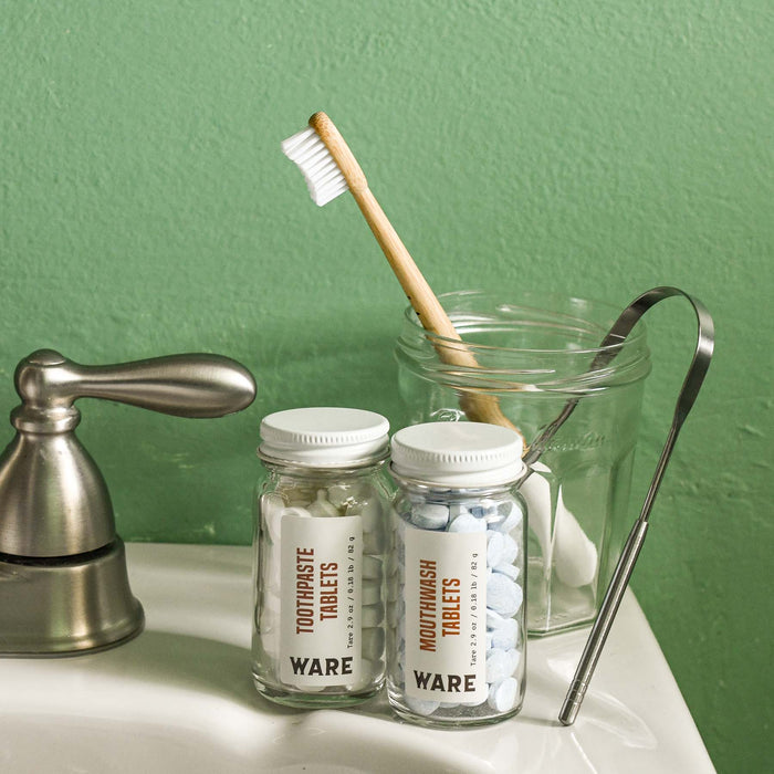 Compostable bamboo toothbrush in bathroom cup with a tongue scraper, toothpaste tablets, and mouthwash tablets.